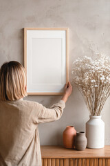 Woman hanging a blank wooden frame on a textured wall in a cozy minimalist interior with vases and dried flowers, ideal for art or poster mockup.