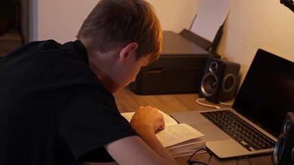 Diligent young student seated at desk in evening at home, deeply engaged in study from textbook, while laptop sits close by, exemplifying dedication to remote learning