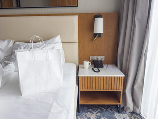 White shopping bag on hotel bed next to bedside table with phone and cup, modern hotel room interior, travel, hospitality and lifestyle concept with copy space