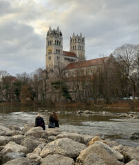 Two women sitting and talking on the shores of the Isar River  in Munich Germany on a winter day with St. Maximilian Catholic Church in the background.