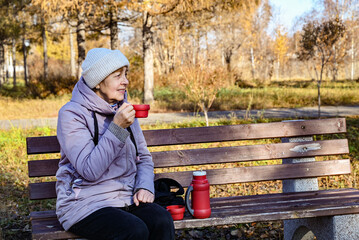 Older woman enjoys warm drink on a park bench during autumn afternoon