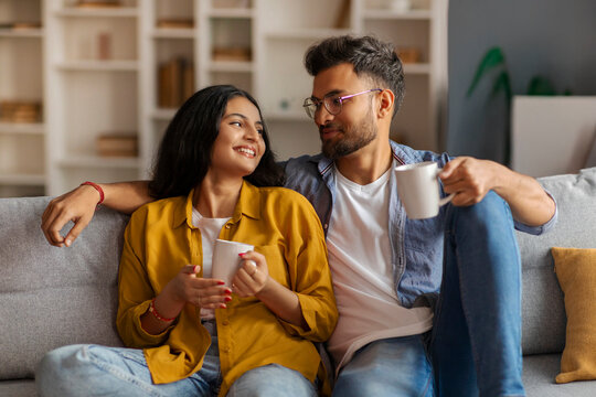 Happy Indian lovers in casual outfits sitting on sofa in living room, drinking tea or coffee, having pleasant conversation, looking at each other and smiling