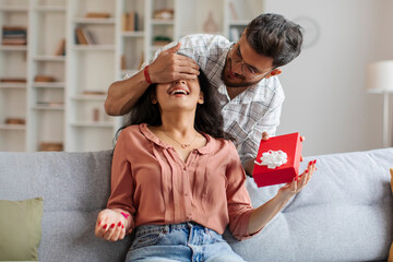 Indian guy closes eyes to surprised woman and giving present in box, lady sitting on couch in living room. Party at home, birthday gift, anniversary, Valentines day and holiday