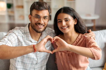 Affectionate Indian couple smiling and creating heart shape together with their hands, symbolizing love and connection, sitting on sofa at home