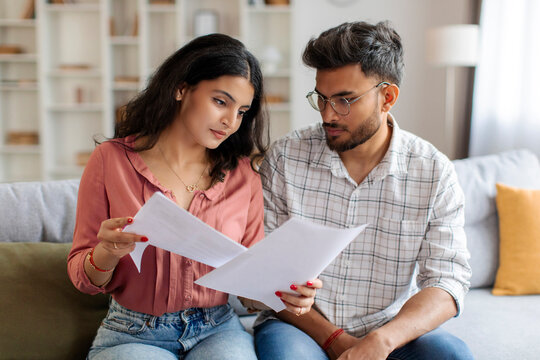 Focused Indian couple checking bills, reading loan documents, counting monthly spendings, planning family budget while woman holding papers