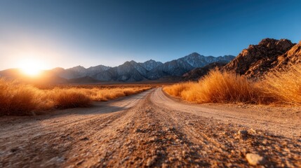 A serene view of a winding dirt road surrounded by golden grasses, leading towards a breathtaking mountain range under a clear blue sky at sunrise, evoking peace and adventure.