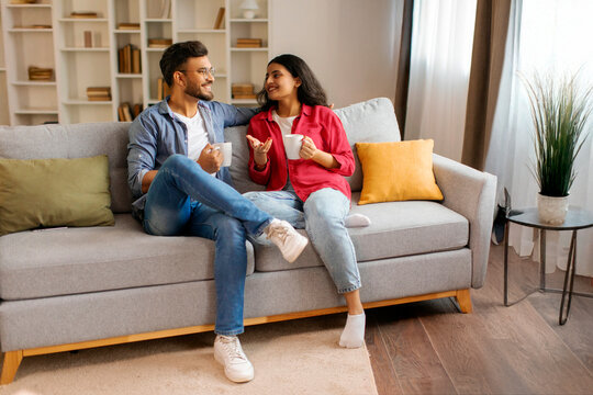Happy Indian couple drinking coffee and having conversation, enjoying weekend together at home, sitting on couch in cozy living room, full length