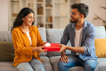 Loving Indian man giving wrapped gift box to his happy wife, celebrating anniversary or birthday, making surprise, sitting together on sofa at home