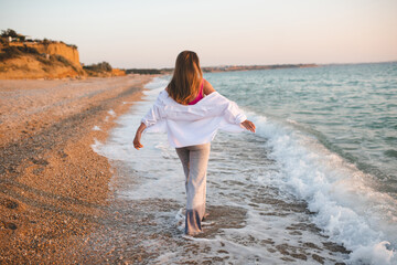 Happy young adult carefree woman running at beach over sea shore wearing white shirt and denim pants. Summer vacation season. Rear view.