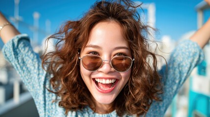 A young woman wearing stylish sunglasses joyfully celebrates under a clear blue sky, embodying happiness and vibrant energy in an upbeat outdoor setting, radiating positivity.