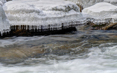 Icicles hang delicately from a snow-covered rock as a stream flows rapidly below. The contrast between the icy formations and the moving water creates a dynamic scene.
