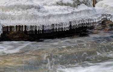 Icicles hang delicately from a snow-covered rock as a stream flows rapidly below. The contrast between the icy formations and the moving water creates a dynamic scene.