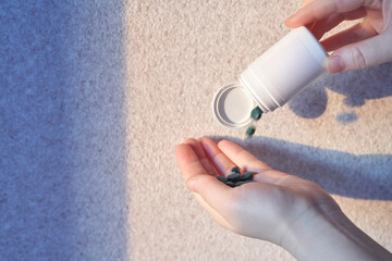 Hands holding white bottle of green pills pouring onto palm on light surface.