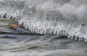 Icicles hang over a flowing river, capturing the contrast between the stillness of the ice and the movement of the water. The scene evokes a sense of tranquility and cold beauty.