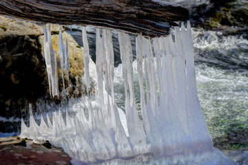 A natural winter scene featuring a horizontal piece of driftwood adorned with icicles of varying lengths and thicknesses. The frozen formations extend downward, merging with ice on nearby rocks 