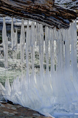 A striking winter scene featuring a fallen tree or log suspended above a flowing stream. Long, slender icicles hang from the textured wood, varying in length 
