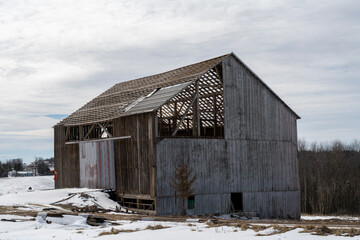 An old wooden barn stands in a rural winter landscape, its roof partially missing and trusses exposed. The aged structure shows signs of disrepair, with damaged siding.