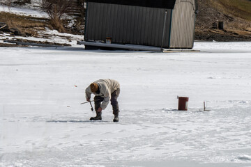 A winter scene capturing a person engaged in ice fishing on a frozen lake. Dressed in warm clothing, the individual bends over near a hole in the ice,