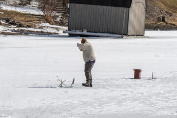 A winter scene capturing a person engaged in ice fishing on a frozen lake. Dressed in warm clothing, the individual bends over near a hole in the ice, handling a fishing rod or tip-up device. 