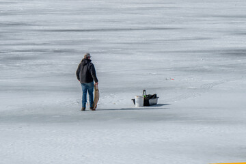 A person walks across a snow-covered frozen lake, carrying a large fish in one hand. Dressed in winter clothing with a cap, jacket, and jeans, the individual moves