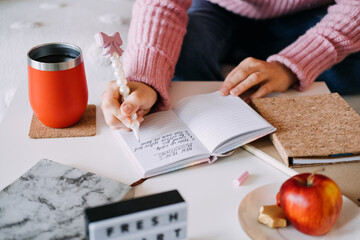 Fototapeta na wymiar Person writes New Year resolutions list in small notebook using pearl pen next to coffee tumbler and apple. New Year resolutions, goal setting, journaling, healthy lifestyle.