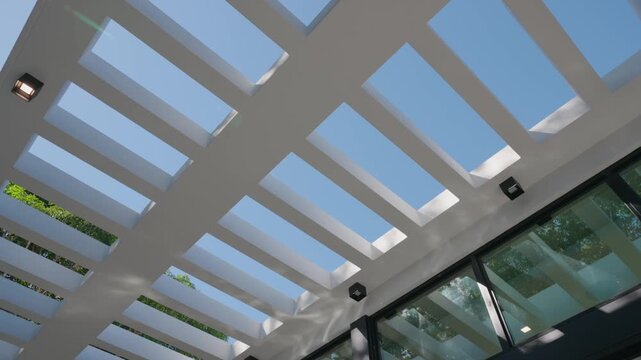 White pergola roof with rectangular cutouts casts shadows on glass walls. Small lights and blue sky add clean, modern feel to open architectural structure.