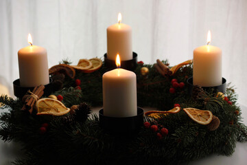 Handmade advent wreath with four burning white candles, spruce branches, dried orange slices, cinnamon sticks and red berries, creating a warm and cozy Christmas atmosphere on a winter table