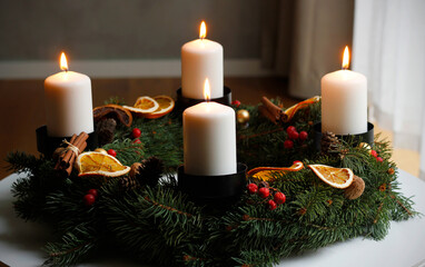 Handmade advent wreath with four burning white candles, spruce branches, dried orange slices, cinnamon sticks and red berries, creating a warm and cozy Christmas atmosphere on a winter table