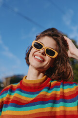 Woman smile sunglasses sweater rainbow portrait happy fashion colorful yellow striped young outdoor sunny blue sky closeup candid model enjoying natural light and cheerful mood.