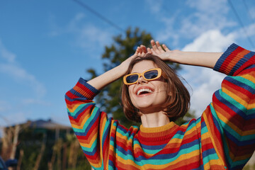 Woman smile sunglasses sweater happy portrait outdoors sunshine in colorful striped jumper, cheerful young adult enjoying a sunny day with hands raised and looking up at sky. © SHOTPRIME STUDIO