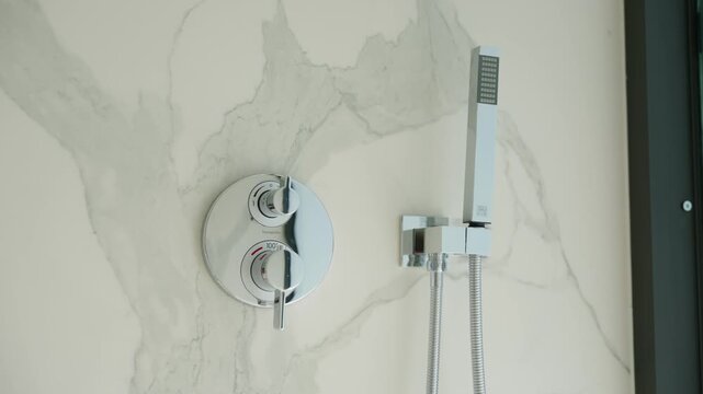Modern shower equipment placed on marble wall. Chrome knobs control water and temperature. Handheld showerhead with metal hose adds clean and simple look.