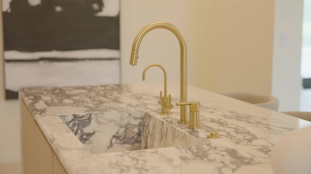 Marble counter with two gold faucets and built in sink creates clean and elegant look. Abstract art and soft tones add cozy feel to modern kitchen space.