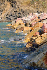 Rocky coastline with colorful stones and gentle waves washing ashore, showcasing rugged seaside textures and clear blue water in natural sunlight.