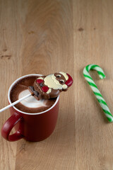 Cozy winter drink setup with a red mug of hot chocolate, a melting chocolate Santa stirrer, and a green and white candy cane on wood