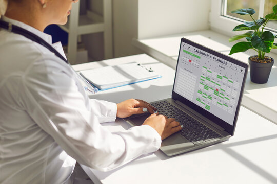 Doctor in office using laptop calendar schedule. Healthcare professional manages online bookings, plans visits, types notes at desk. Medical scheduling and telehealth productivity concept.
