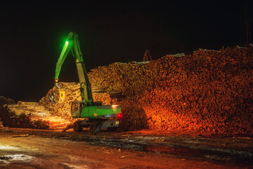 Night operation at an industrial port terminal where a material handler crane stacks and moves large piles of timber logs. The scene shows wood cargo handling under artificial lighting