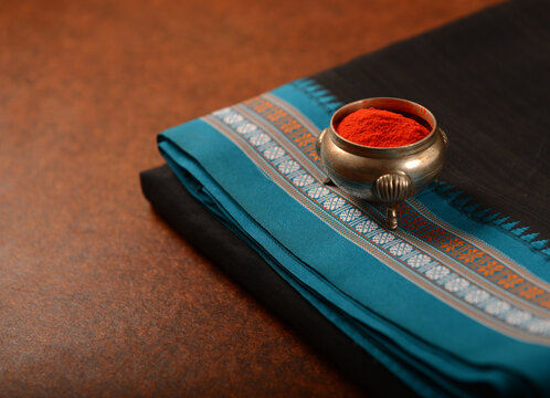 An ornate silver bowl filled with vibrant red Kumkum powder rests on a folded black saree with detailed teal boarder. A Makar sankranti festive objects and background.