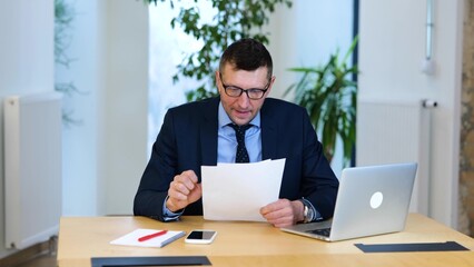 Fototapeta premium video. Caucasian businessman in black suit and blue shirt is sitting at the table, working on his computer, reading documents, and feels success.