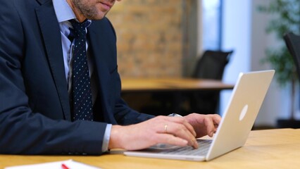 Caucasian man in a black suit and blue shirt and glasses is sitting and typing on his computer at a working table. Slow-motion video, close up.