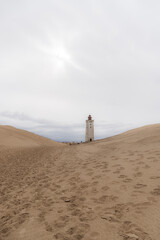 tall, weathered lighthouse, barren sand dunes, pale, overcast sky, minimalist, atmospheric coastal scene. olitude, timelessness, and navigation, travel, resilience, and remote landscapes., backlit