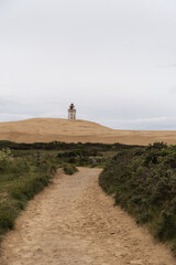 sandy footpath winding through coastal vegetation toward solitary lighthouse atop golden sand dunes, set against overcast sky, calm, atmospheric coastal scene. green vegetation bushes in forgreground