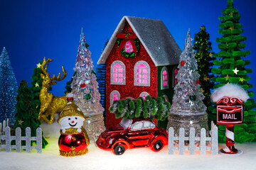 A snowman and a glass Christmas tree with a red car near a red Christmas house surrounded by a fence