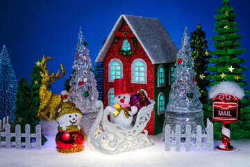 A snowman and a snowman in a red hat on a white sled with a gift near a red Christmas house surrounded by a fence