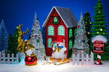 A snowman and a snowman on a sled among glass Christmas trees near a red Christmas house surrounded by a fence