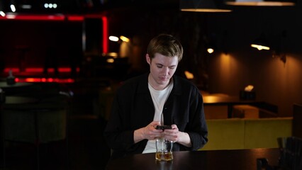 Upset young man reading message while holding mobile phone, drinking whiskey, sitting in the...