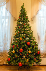 Christmas tree adorned with colorful ornaments and golden decorations stands elegantly in a bright room with soft drapes, creating a festive holiday atmosphere
