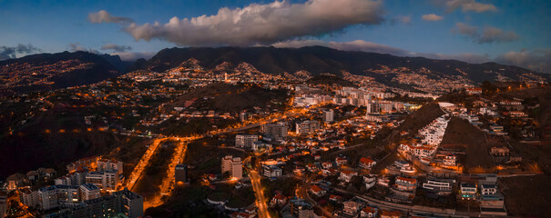 Evening view of Funchal, Madeira Island, with winding lit roads, terraced houses, and mid rise blocks rising toward dark ridges, long exposure glow, cool blue dusk.