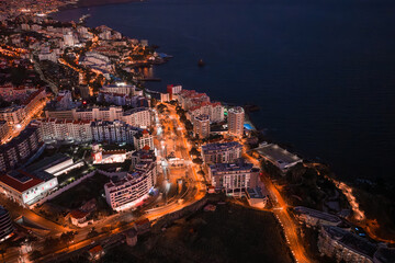 Aerial twilight view of Funchal, Madeira shows terraced white buildings, a curving waterfront road...