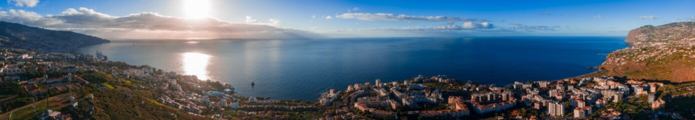 Wide angle view of Funchal, Portugal, terraced hills and white roofs by the Atlantic, late afternoon light, calm water, rugged cliffs framing the bay, long reflections.