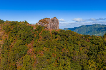 Aerial view shows a forested ridge with a rocky outcrop on Madeira Island, Portugal. Distant mountains frame the horizon in late summer light with russet and green tones.